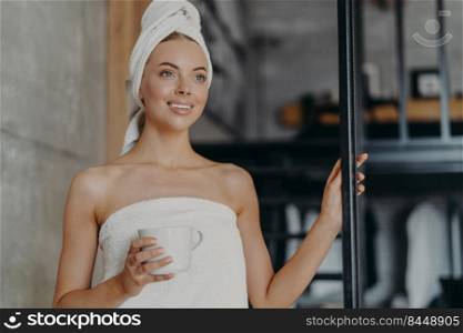 Beautiful young female looks thoughtfully into distance, drinks coffee or tea, poses with bare shoulders, wrapped in bath towel after showering, smiles pleasantly, has white teeth. Skin care concept