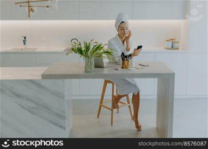 Beautiful young female model poses in modern apartment wears bathrobe and towel on head applies facial cream undergoes beauty procedures uses cosmetic tools for skin care. P&ering wellness