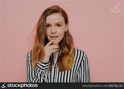 Beautiful young female student dressed in striped shirt touching chin and can not believe her eyes, looking at camera with amazed face expression, posing on pink background in studio. Excited young woman with open mouth holding hand on chin
