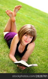 beautiful young female student reading a book outdoors laying on grass