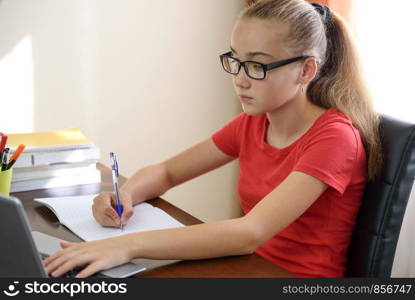 Beautiful young female student with glasses doing homework on a laptop and a notebook in her room.