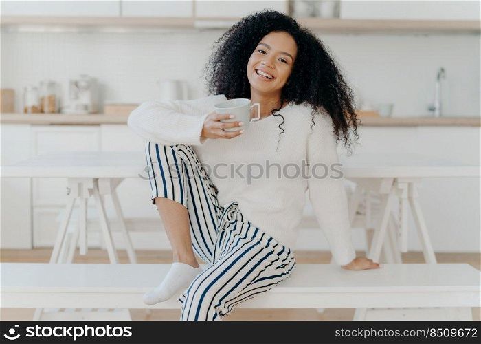 Beautiful young female with Afro haircut, poses on white bench indoor, dressed in stylish clothing, enjoys aromatic beverage, enjoys breakfast at home, poses against blurred kitchen interior
