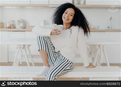 Beautiful young female with Afro haircut, poses on white bench indoor, dressed in stylish clothing, enjoys aromatic beverage, enjoys breakfast at home, poses against blurred kitchen interior