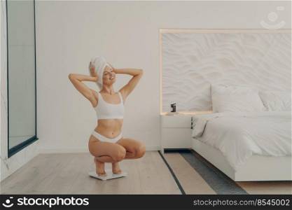 Beautiful young fit female model squats on scales in modern bedroom interior after morning shower routine in underwear happy and satisfied with result of her weight. Weight loss and dieting concept. Beautiful young fit female model squats on scales in modern bedroom interior