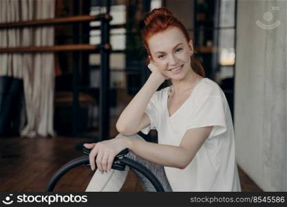 Beautiful young ginger female fitness instructor in white tshirt sitting on floor and smiling on camera while perfoming pilates ring workout in gym. Concept of healthy and active lifestyle. Satisfied young woman in sportswear with pilates ring