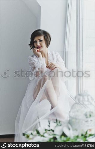Beautiful young girl in a transparent negligee sitting on a window sill with flowers.. Portrait of a girl in a negligee on background of the window 6388.