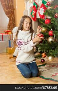 Beautiful young girl in sweater sitting on floor at living room and decorating Christmas tree