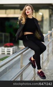 Beautiful young girl wearing black jacket sitting in the street.. Young blonde girl with beautiful blue eyes wearing black jacket and trousers outdoors. Pretty russian female with long wavy hair hairstyle. Woman in urban background.