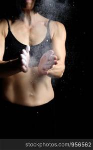 beautiful young girl with a sports figure dressed in a black top claps in her hands with white magnesia, preparing before exercise, dark key