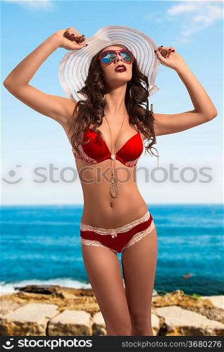 beautiful young girl with brown hair in fashion pose wearing red sexy bikini, white hat and necklace close to sea