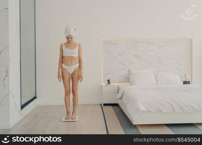 Beautiful young good looking female in underwear with head wrapped in towel looking down while standing in bedroom on scale and measuring her weight. Beauty and body care concept. Beautiful young sportive female in underwear with head wrapped in towel measuring her weight