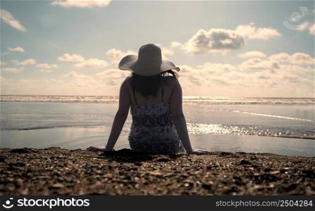 Beautiful young Hispanic woman sitting alone at the edge of the beach seen from behind, wearing a hat and a black and white dress during sunset. Beautiful young Hispanic woman sitting alone at the edge of the beach wearing a hat and a black and white dress during sunset