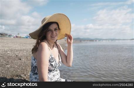 Beautiful young Hispanic woman smiling sitting alone at the edge of the beach looking at the camera, holding her hat with her hand, wearing a black and white dress during a sunny morning. Beautiful happy young Hispanic woman sitting alone at the edge of the beach looking at the camera, wearing a hat and a black and white dress during a sunny morning