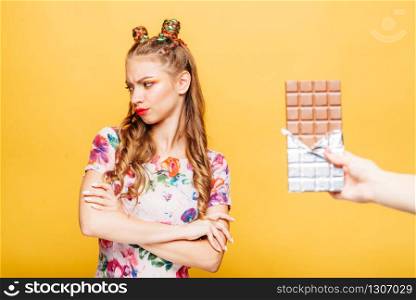Beautiful young lady refuses to take a chocolate bar. Bright girl with blonde curly hair. Stylish girl in summer colorful dress, yellow wall on background.. Lady refuses to take a chocolate bar
