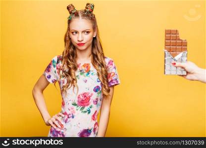 Beautiful young lady refuses to take a chocolate bar. Bright girl with blonde curly hair. Stylish girl in summer colorful dress, yellow wall on background.. Lady refuses to take a chocolate bar