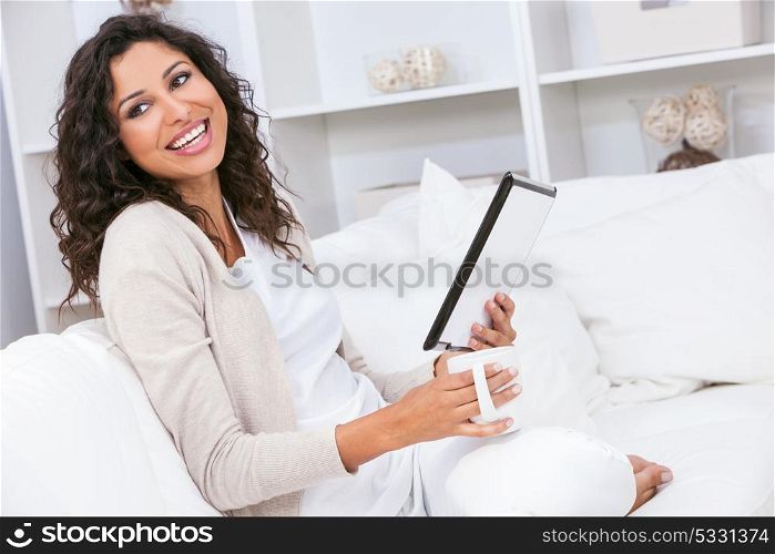 Beautiful young Latina Hispanic woman laughing, smiling, relaxing and drinking a cup of coffee or tea at home using tablet computer