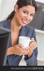 Beautiful young Latina Hispanic woman or businesswoman in smart business suit sitting at a desk drinking coffee in an office