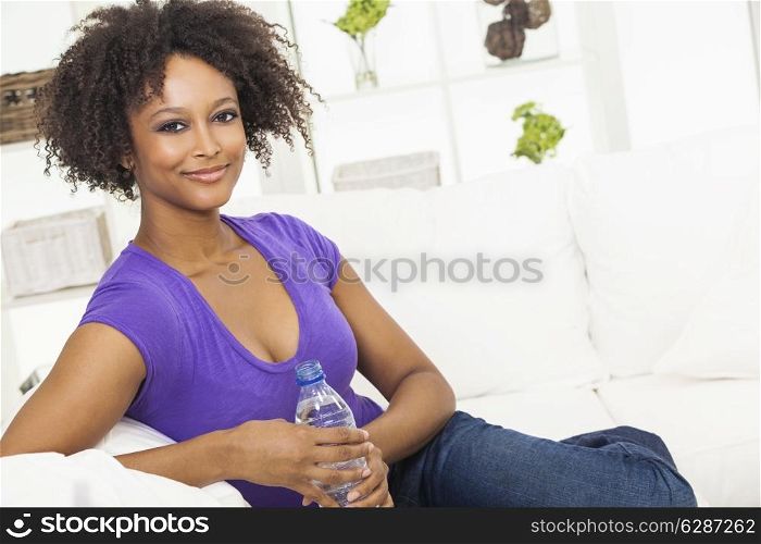 Beautiful young mixed race black African American woman smiling, relaxing and drinking a bottle of water at home on a sofa