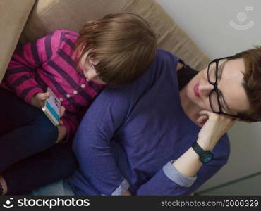 Beautiful young mother and her cute little daughter are using a mobile phone and smiling, sitting on sofa at home