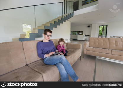 Beautiful young mother and her cute little daughter are using a tablet and smiling, sitting on sofa at home