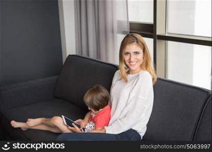 Beautiful young mother and her cute little daughter are using a tablet and smiling, sitting on sofa at home