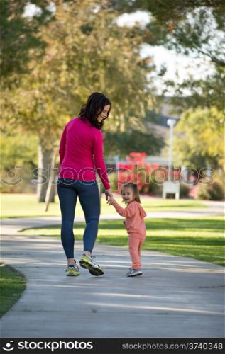 Beautiful young mother and her daughter walking in the neighborhood. They are on a sidewalk in a grassy greenbelt. They are holding hands.