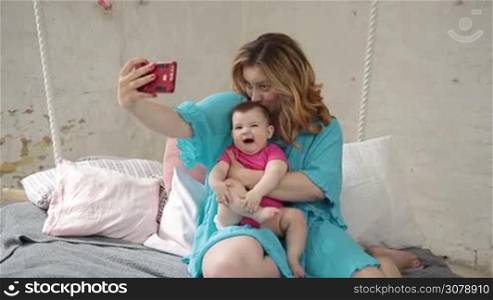 Beautiful young mother emabracing her smiling baby girl and taking selfie with smartphone while enjoying time together at home. Young charming mother and adorable infant child making self-portrait with mobile phone. Front view. Slow motion.