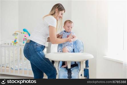 Beautiful young mother seating her baby boy in high chair