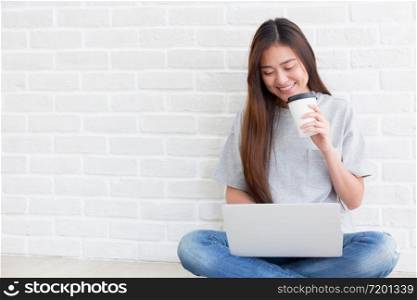 Beautiful young of Asian girl using a laptop while sitting at the floor in the living room and hold coffee, Asia women smile while typing work from home