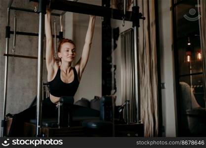 Beautiful young pilates trainer in black sportswear with athletic figure doing full body stretching exercise on cadillac reformer in fitness studio. Concept of healthy lifestyle and sport indoors. Beautiful young ginger woman doing upper body stretching exercise on reformer