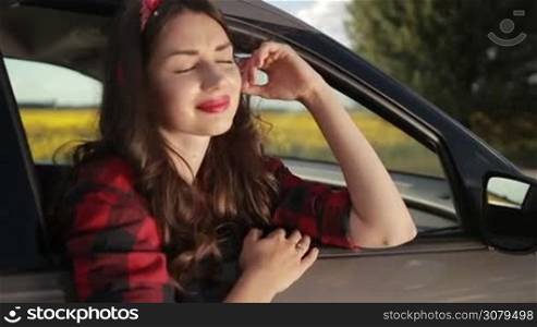 Beautiful young pin-up female wearing red lipstick, headband and plaid shirt sitting in the car, looking out of open window with innocent and radiant smile while enjoying leisure during summer roadtrip vacation.