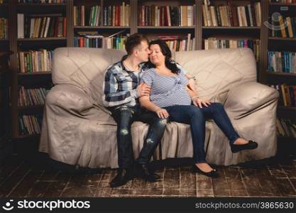 Beautiful young pregnant couple hugging on sofa against bookshelves
