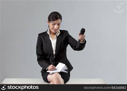 Beautiful young reporter asking comment while writing in notepad against colored background