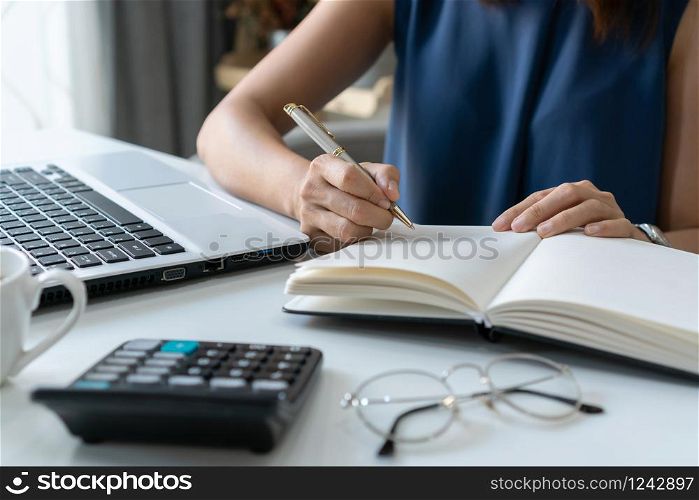 Beautiful young smiling Asian businesswoman writing notebook document finance and calculator at home office.