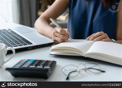 Beautiful young smiling Asian businesswoman writing notebook document finance and calculator at home office.