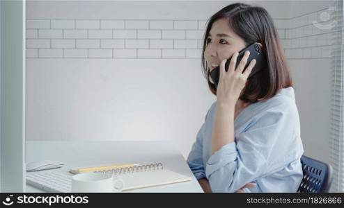 Beautiful young smiling asian woman working on laptop while sitting in a living room at home. Asian business woman using phone for work in her home office. Enjoying time at home.