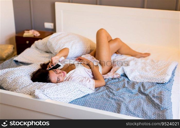 Beautiful young smiling brunette woman lying in bed and using a phone in her bedroom.