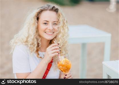 Beautiful young smiling female with curly light hair, toothy smile, recreats on beach, drinks summer fresh cocktail, dressed in casual white t shirt, has good rest. People, summer, lifestyle concept