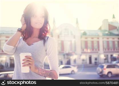 Beautiful young smiling woman with tattoo, holding a disposable takeaway cup and looking aside against urban city background.