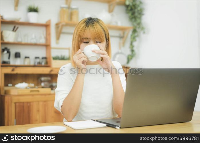 Beautiful young smiling woman working on laptop while enjoying drinking warm coffee sitting in a living room at home. Enjoying time at home. Asian business woman working in her home office.