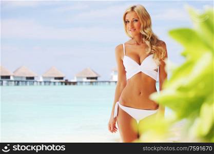 Beautiful young tan woman in white bikini on tropical beach at Maldives villa houses and blue sea on background. Woman on tropical beach