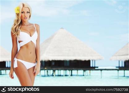 Beautiful young tan woman in white bikini on tropical beach at Maldives villa houses and blue sea on background. Woman on tropical beach