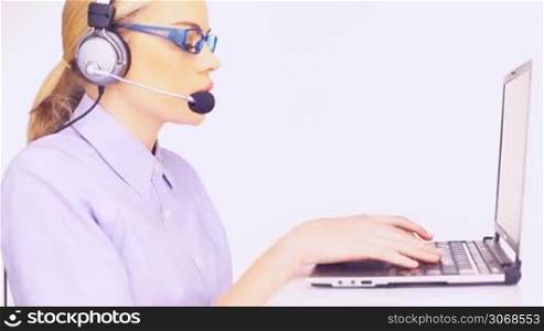 Beautiful young telesales lady wearing glasses and and a headset working at her laptop computer turning to smile at the camera