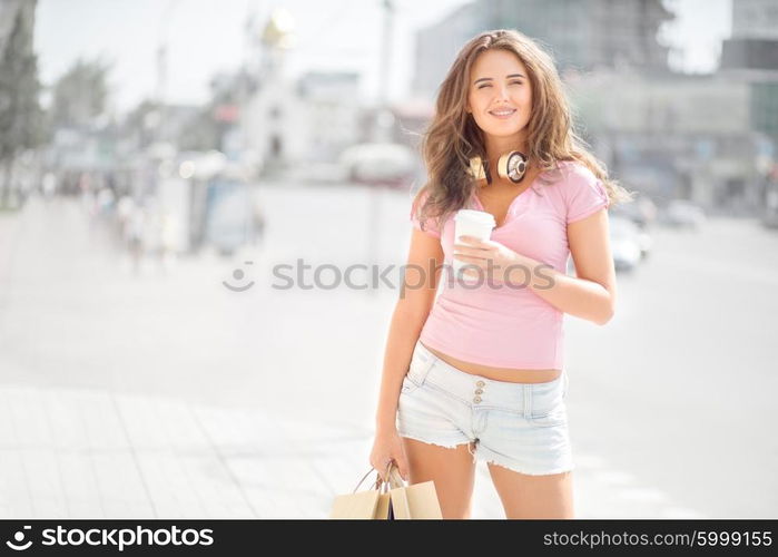 Beautiful young trendy woman wearing music headphones around her neck, drinking take away coffee and walking with shopping bags in an urban city.