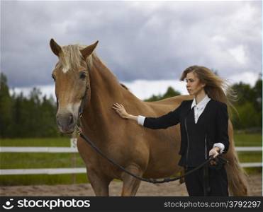 Beautiful young woman and a brown stallion, cloudy sky and rural landscape on background