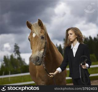 Beautiful young woman and a brown stallion, cloudy sky and rural landscape on background