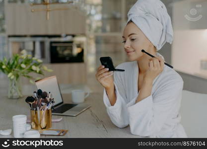 Beautiful young woman applies makeup cosmetics with brush in front of mirror poses against home interior sits at desk with opened laptop and cosmetology tools dressed in bathrobe. Self care concept