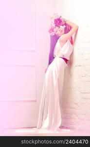 Beautiful young woman / bride in white long dress holding bouquet of peonies and roses lean against the wall