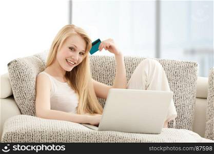 Beautiful Young Woman Doing Online Shopping onl Laptop with a Credit Card, Sitting on the Sofa in Living Room
