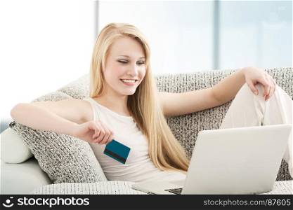 Beautiful Young Woman Doing Online Shopping onl Laptop with a Credit Card, Sitting on the Sofa in Living Room
